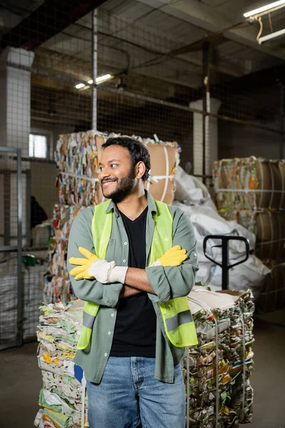 Positive indian sorter in protective gloves and vest crossing arms while standing near waste paper on hand pallet truck in blurred waste disposal station, garbage sorting and recycling concept — Stock Photo