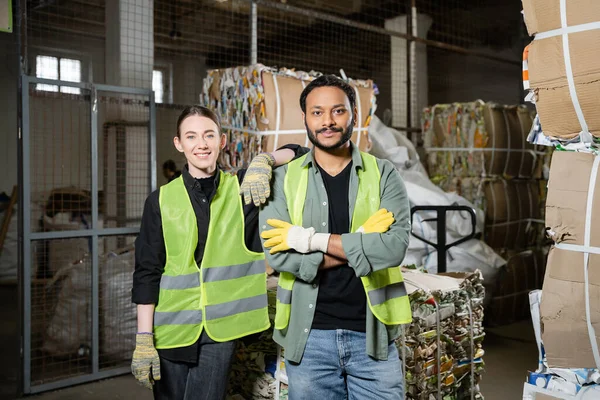 Smiling young worker in protective vest and gloves looking at camera near indian colleague crossing arms and standing near waste paper in waste disposal station, garbage sorting and recycling concept — Stock Photo