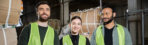 Cheerful and interracial workers in high visibility vests looking at camera while standing near waste paper in blurred waste disposal station, garbage sorting and recycling concept, banner — Stock Photo
