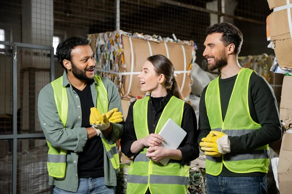 Smiling indian worker in safety vest and gloves talking to colleagues with digital tablet while standing near blurred waste paper in waste disposal station, garbage sorting and recycling concept — Stock Photo
