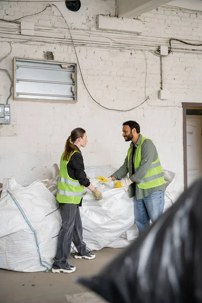 Side view of indian worker in protective vest and gloves standing near young colleague and sacks while working in waste disposal station, garbage sorting and recycling concept — Stock Photo