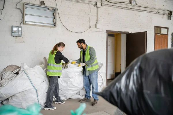 Seitenansicht eines jungen Arbeiters in Warnweste und Handschuhen, der neben Säcken und einem indischen Kollegen steht, während er in der Müllentsorgungsstation, im Müllsortier- und Recyclingkonzept arbeitet — Stockfoto