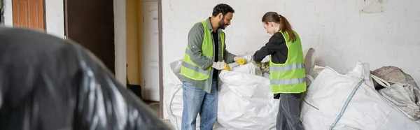Vista lateral de trabalhadores multiétnicos em coletes de proteção e luvas perto de sacos e trabalhando em estação de eliminação de resíduos, triagem de lixo e conceito de reciclagem, bandeira — Fotografia de Stock