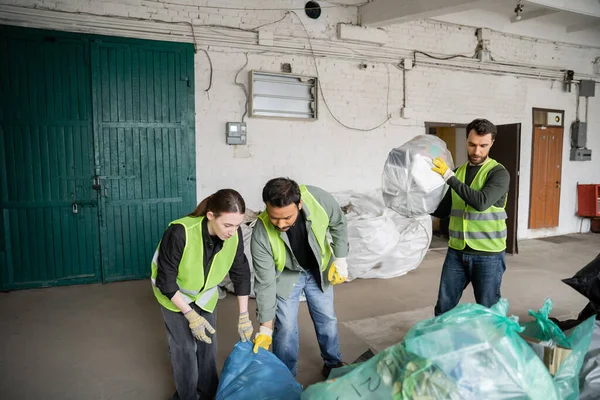 Trabajadores multiétnicos en chalecos y guantes de protección que trabajan con bolsas de plástico cerca de su colega en la estación de eliminación de residuos, clasificación de basura y el concepto de reciclaje - foto de stock