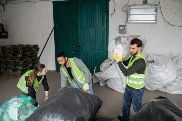 Trabajadores multiétnicos en chalecos y guantes de alta visibilidad que trabajan con bolsas de plástico cerca de su colega en la estación de eliminación de residuos, clasificación de basura y el concepto de reciclaje - foto de stock