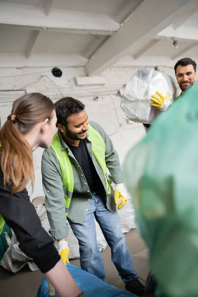 Улыбающийся индийский рабочий в бронежилете и перчатках, работающий рядом с коллегами и пластиковыми пакетами на станции утилизации отходов, концепция сортировки и переработки мусора — стоковое фото