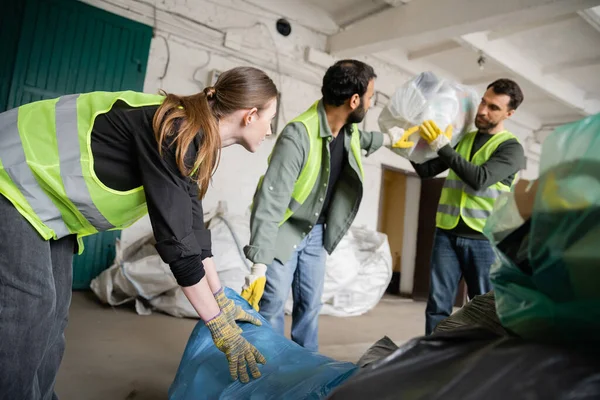 Jovem trabalhadora em colete de proteção e luvas segurando saco plástico perto de colegas multiétnicos borrados na estação de descarte de resíduos, conceito de triagem e reciclagem de lixo — Fotografia de Stock