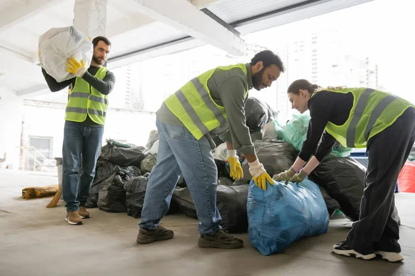 Улыбающаяся работница в бронежилете и перчатках, выкладывающая пластиковый пакет с мусором рядом с многонациональными коллегами в мусоросборник, концепция сортировки и переработки мусора — стоковое фото