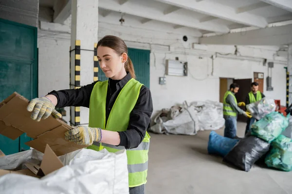 Young female worker in protective vest and gloves holding cardboard near sack and blurred colleagues working in waste disposal station, garbage sorting and recycling concept — Stock Photo