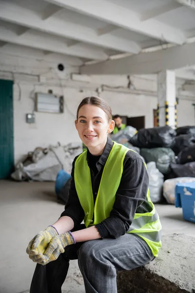 Positive young worker in protective vest and gloves looking at camera while resting near blurred plastic bags with trash at background in garbage sorting center, recycling concept — Stock Photo