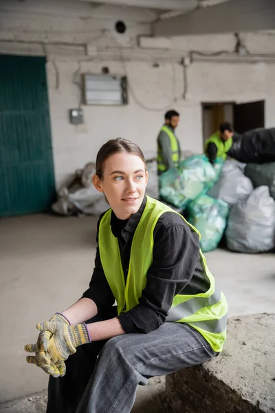 Positive junge Sortierer in Warnweste und Handschuhen, die wegschauen, während sie sich in der Nähe verschwommener Kollegen und Plastiktüten im Hintergrund im Müllsortierzentrum entspannen, Recyclingkonzept — Stockfoto
