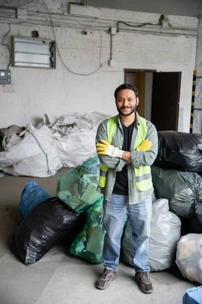 Fröhlicher und bärtiger indischer Arbeiter in Schutzweste und Handschuhen, der die Arme verschränkt und in die Kamera blickt, während er in der Müllsortieranlage neben Plastiktüten steht, Recyclingkonzept — Stockfoto