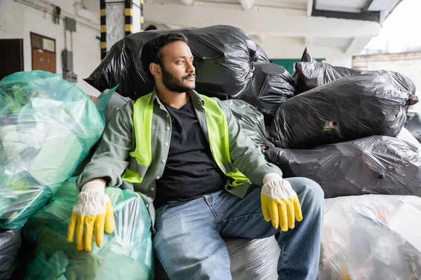 Bärtiger indischer Sortierer in fluoreszierender Weste und Schutzhandschuhen, der wegschaut, während er auf Plastiktüten mit Müll im Müllsortierzentrum sitzt, Recyclingkonzept — Stockfoto