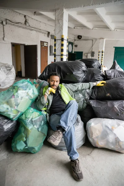 Fiducioso lavoratore indiano in giubbotto e guanti ad alta visibilità guardando la fotocamera mentre seduto su sacchetti di plastica con immondizia nel centro di smistamento della spazzatura, concetto di riciclaggio — Foto stock