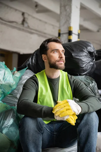 Trabajador barbudo en chaleco de alta visibilidad y guantes mirando hacia otro lado mientras está sentado cerca de bolsas de plástico con basura en el centro de clasificación de basura borrosa, concepto de reciclaje - foto de stock