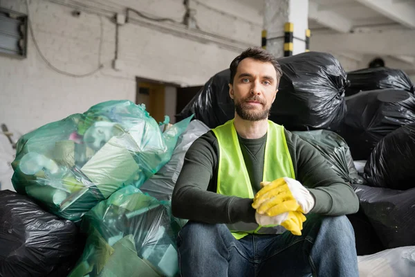 Bärtiger Sortierer in Schutzweste und Handschuhen, der in die Kamera blickt, während er in der Nähe von Plastiktüten mit Müll sitzt, während er in einem verschwommenen Müllsortierzentrum arbeitet, Recyclingkonzept — Stockfoto