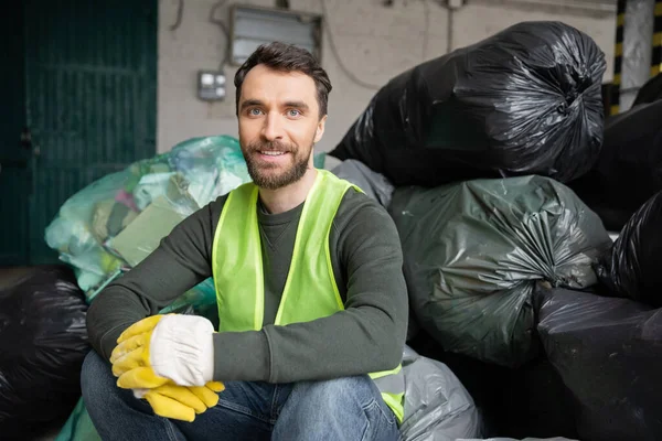 Lächelnder und bärtiger Arbeiter in Warnweste und Handschuhen, der in die Kamera blickt, während er auf Plastiktüten mit Müll in verschwommenem Müllsortierzentrum sitzt, Recyclingkonzept — Stockfoto