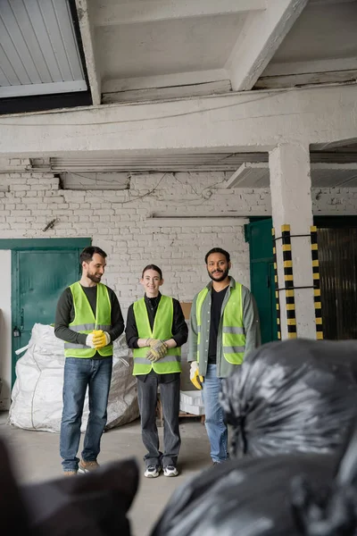 Positive multiethnic sorters in protective gloves and vests looking at camera while standing near blurred plastic bags with trash in garbage sorting center, recycling concept — Stock Photo