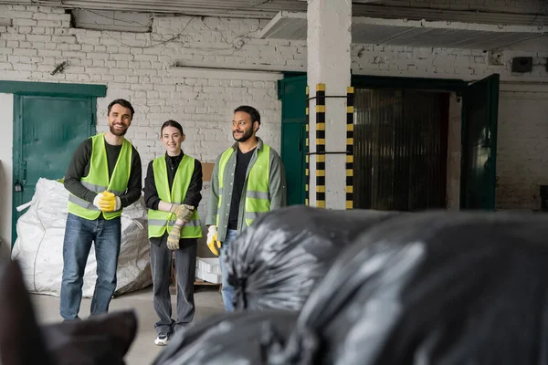 Trabajadores multiétnicos positivos en chalecos fluorescentes y guantes protectores mirando a la cámara mientras están de pie cerca de bolsas de plástico borrosas en el centro de clasificación de basura, concepto de reciclaje - foto de stock