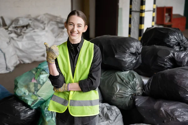 Lächelnder junger Arbeiter in Schutzweste und Handschuhen, der wie eine Geste wirkt und in die Kamera schaut, während er neben verschwommenen Plastiktüten mit Müll im Müllsortierzentrum steht, Recyclingkonzept — Stockfoto