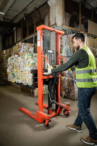 Side view of bearded sorter in protective vest and gloves using hand pallet truck with waste paper while working in blurred garbage sorting center, recycling concept — Stock Photo