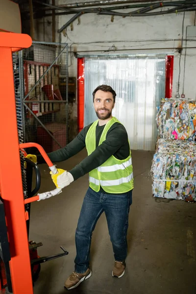 Trabajador sonriente en chaleco de seguridad y guantes tirando de la transpaleta de mano mientras trabaja cerca de papel de desecho borroso en el centro de clasificación de basura en segundo plano, concepto de reciclaje - foto de stock
