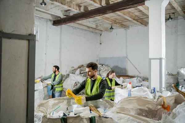 Male worker in protective vest and gloves holding plastic trash while standing near sacks and blurred interracial colleagues in waste disposal station, garbage sorting and recycling concept — Stock Photo