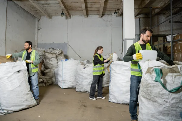Trabajadores multiétnicos en chalecos y guantes de alta visibilidad que separan la basura en sacos mientras trabajan en la estación de eliminación de residuos, la clasificación de basura y el concepto de reciclaje - foto de stock