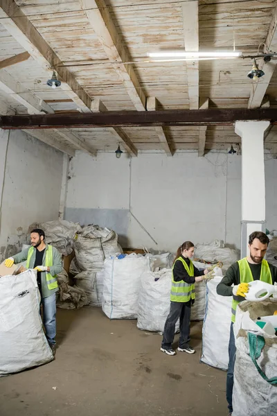 Interracial workers in high visibility vests and gloves soring trash in sacks while working together in waste disposal station, garbage sorting and recycling concept — Stock Photo