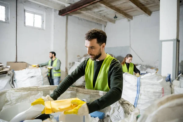 Bearded worker in protective gloves and vest taking plastic trash from sack while working near blurred colleagues in waste disposal station, garbage sorting and recycling concept — Stock Photo