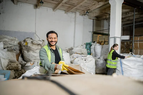 Cheerful indian worker in protective vest and gloves looking at camera while sorting trash near sacks and colleague in waste disposal station, garbage sorting and recycling concept — Stock Photo