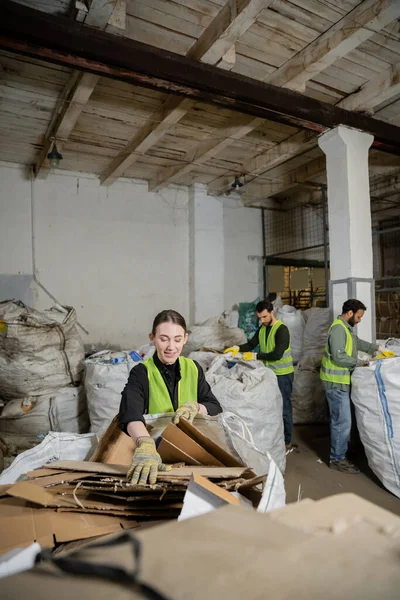 Trabalhador positivo em luvas e colete colocando papelão no saco enquanto separa o lixo perto de colegas multiétnicos borrados na estação de eliminação de resíduos, triagem de lixo e conceito de reciclagem — Fotografia de Stock