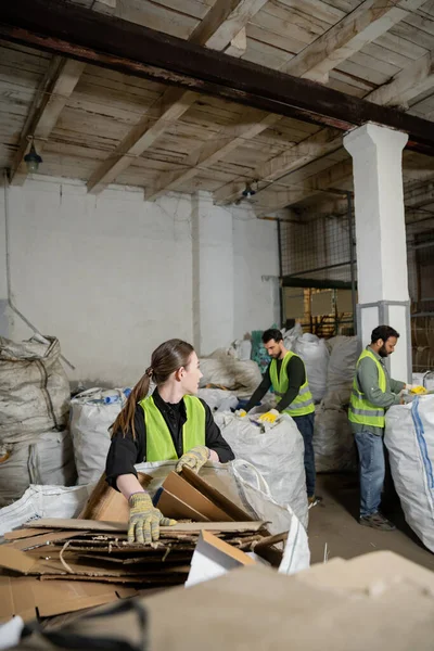 Junge Arbeiterinnen in Schutzhandschuhen und Weste schauen multiethnische Kollegen an, während sie Karton in Säcke in Mülldeponie, Müllsortier- und Recyclingkonzept packen — Stockfoto