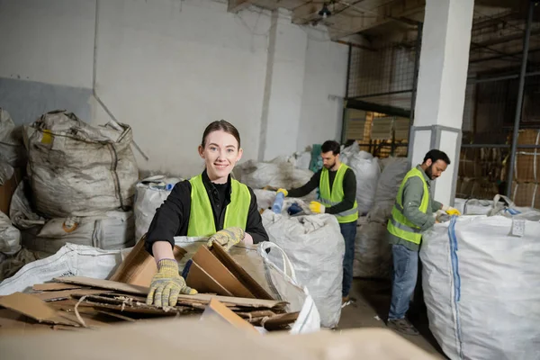 Worker in gloves and high visibility vest looking at camera while putting cardboard in sack and working near multiethnic colleagues in waste disposal station, garbage sorting and recycling concept — Stock Photo