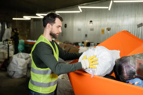 Side view of sorter in glove and protective vest putting sack in container while working in blurred waste disposal station at background, garbage sorting and recycling concept — Stock Photo
