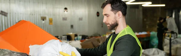 Side view of bearded sorter in protective vest and glove putting trash sack in container in blurred waste disposal station, garbage sorting and recycling concept, banner — Stock Photo