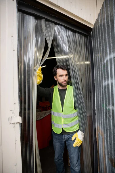 Бородатий чоловік-працівник у жилеті та рукавичках високої видимості, що стоїть біля входу на станцію утилізації відходів, концепція сортування сміття та переробки — стокове фото