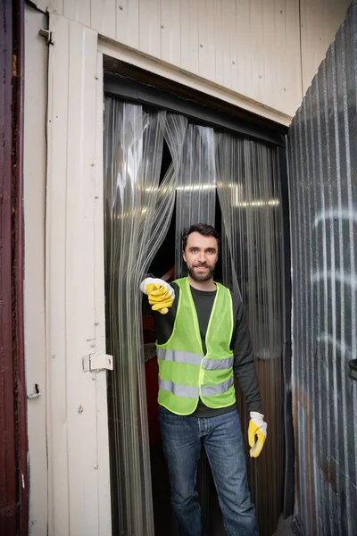Trabalhador masculino alegre em colete fluorescente e luvas de proteção olhando para a câmera enquanto está perto da porta da estação de eliminação de resíduos, triagem de lixo e conceito de reciclagem — Fotografia de Stock