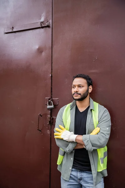 Der bärtige indische Arbeiter in Warnweste und Handschuhen kreuzt die Arme, während er in der Nähe der Tür der Mülldeponie steht, Müllsortier- und Recyclingkonzept — Stockfoto