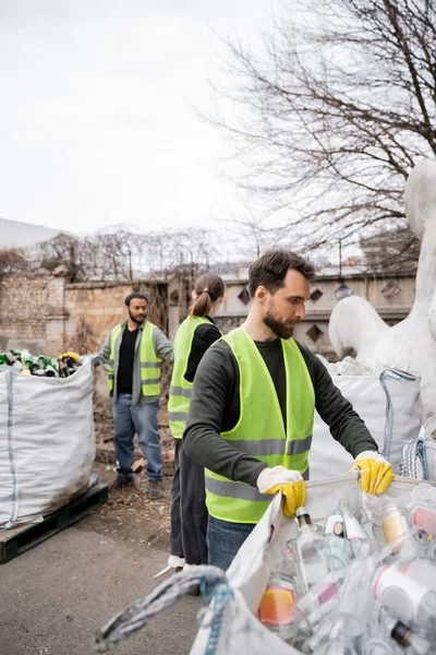 Bearded worker in safety vest and gloves holding sack with glass trash near blurred multiethnic colleagues in outdoors waste disposal station, garbage sorting and recycling concept — Stock Photo