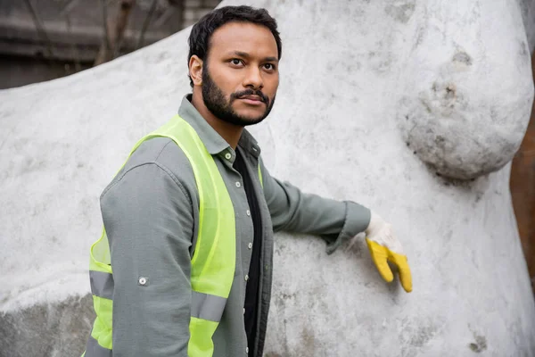 Серйозний індійський працівник у жилеті та рукавичці, що стоїть біля бетонної скульптури на станції утилізації зовнішніх відходів, концепція сортування сміття та переробки — стокове фото