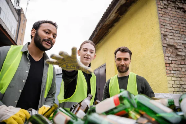 Trabalhadores multiétnicos sorridentes em luvas e coletes de proteção olhando para lixo de vidro desfocado na estação de eliminação de resíduos ao ar livre, triagem de lixo e conceito de reciclagem — Fotografia de Stock