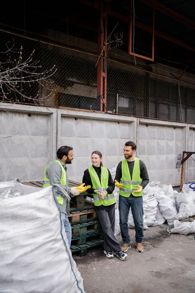 Lächelnder indischer Arbeiter in Warnweste und Handschuhen, der sich mit fröhlichen Kollegen unterhält, während er neben Säcken mit Müll in der Müllentsorgungsstation steht, Mülltrennung und Recyclingkonzept — Stockfoto
