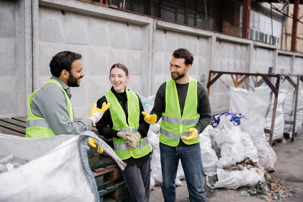 Trabalhador positivo em colete de alta visibilidade e luvas conversando com colegas multiétnicos perto de sacos com lixo em estação de descarte de resíduos ao ar livre, conceito de triagem e reciclagem de lixo — Fotografia de Stock