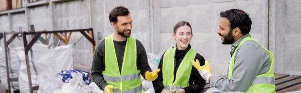 Cheerful multiethnic sorters in high visibility vests and gloves talking while standing near trash in outdoor waste disposal station, garbage sorting and recycling concept, banner — Stock Photo