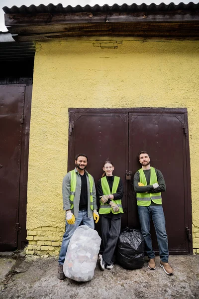 Trabalhadores inter-raciais sorridentes em luvas e coletes de proteção olhando para a câmera enquanto estão perto de sacos de plástico com lixo perto da porta da estação de eliminação de resíduos ao ar livre, conceito de triagem de lixo — Fotografia de Stock