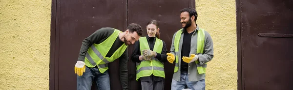 Positive multiethnic workers in high visibility vests and gloves talking while standing near door of waste disposal station outdoors, garbage sorting and recycling concept, banner — Stock Photo