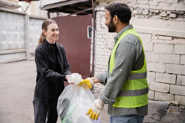 Smiling young volunteer giving plastic bag with trash to indian worker in protective vest and gloves outdoors in waste disposal station, garbage sorting and recycling concept — Stock Photo