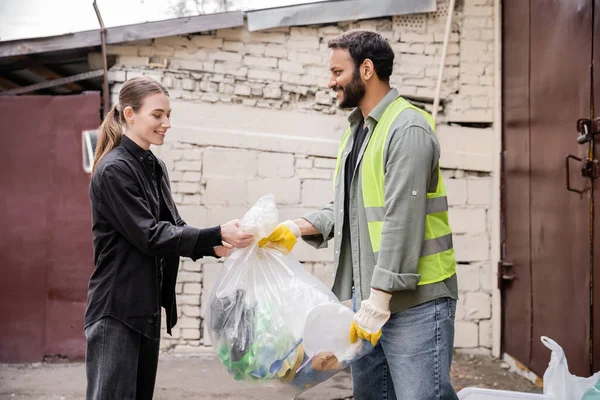 Positive indian worker in safety vest and gloves taking plastic bag with trash from volunteer outdoors in waste disposal station, garbage sorting and recycling concept — Stock Photo