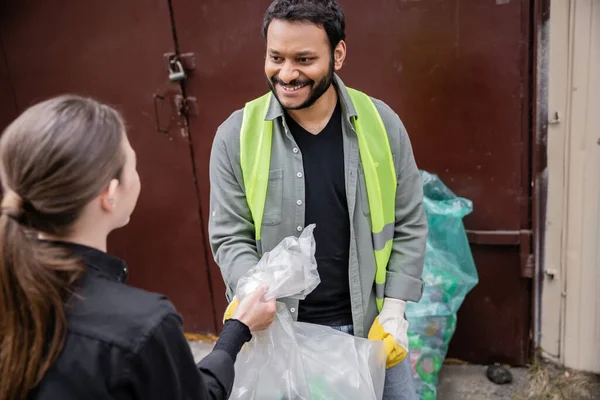 Trabajador indio sonriente en chaleco de seguridad y guantes que toma la bolsa de basura de voluntarios borrosos en la estación de eliminación de residuos al aire libre, clasificación de basura y concepto de reciclaje - foto de stock